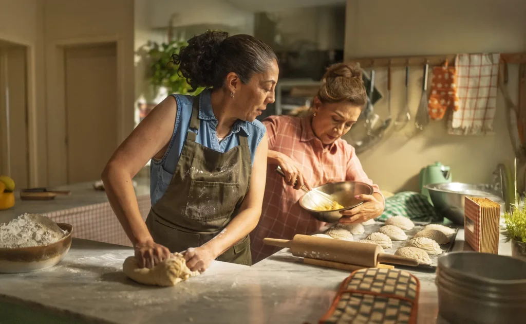 Two women baking together in bright kitchen, kneading dough on floured counter with rolling pin and ingredients